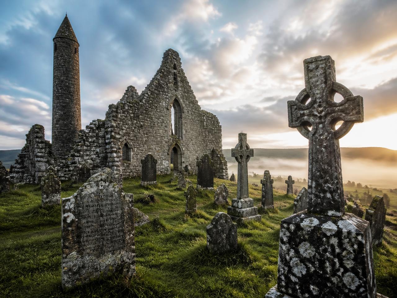 Early Irish monastic ruin with a round tower and high crosses in mist at dawn